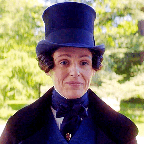 Anne Lister, dressed in a top hat, overcoat, neck kerchief, and vest, looks directly at the camera, raises her eyebrows, smiles, and then begins to move forward. 