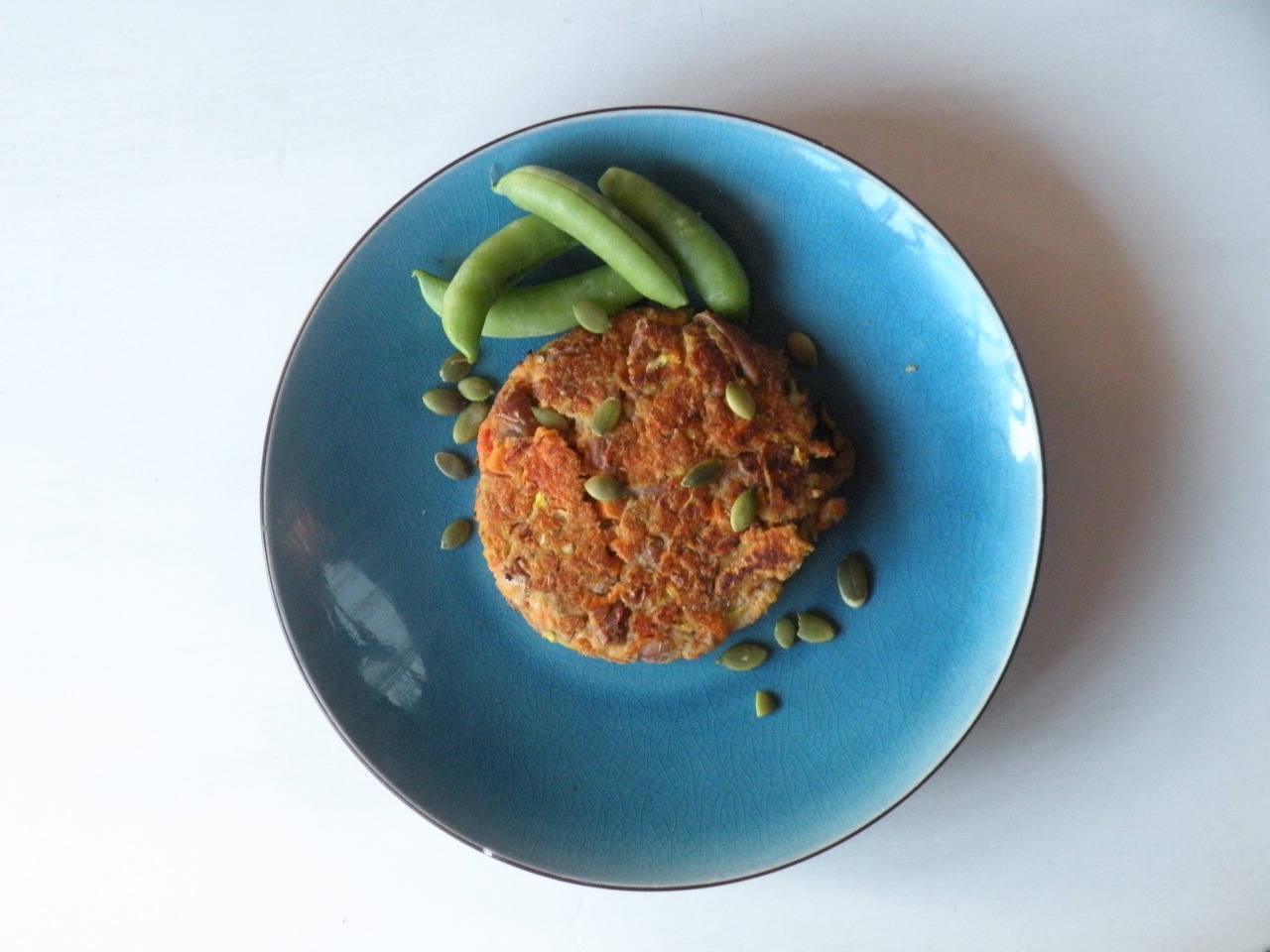 A bean burger on a blue plate, with a small pile of peas next to it and pumpkin seeds scattered on top.