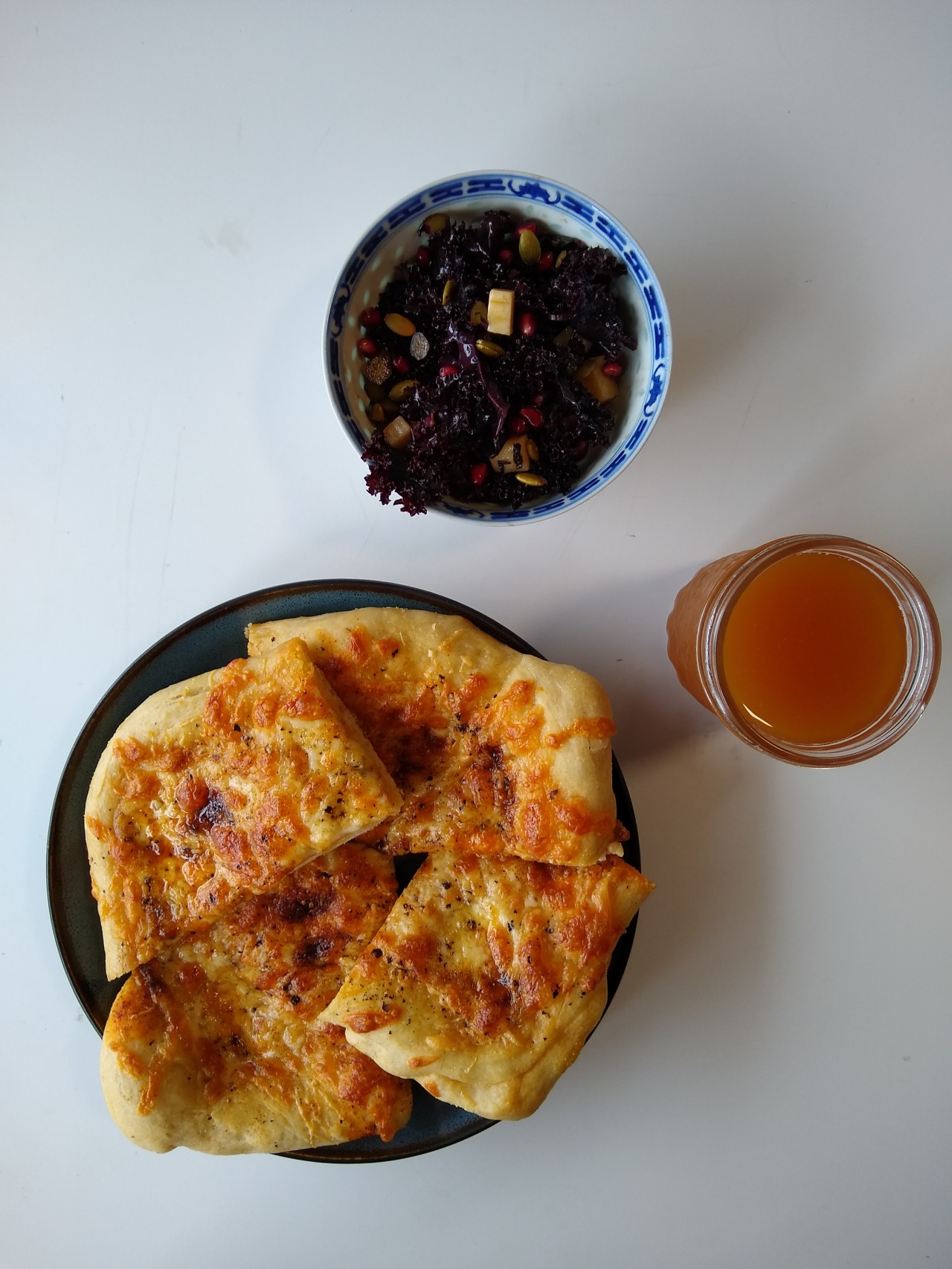 An overhead shot of cheese pizza on a blue plate, purple kale salad in a white and blue bowl, and apple cider in a canning jar, all on a white table.