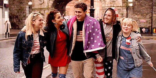 Five characters, four girls one men walking down a street with their arms around each other draped in miss-coloured American flags