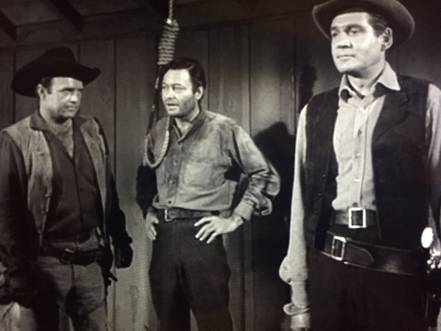 DeForest Kelley stands in a doorway with a very sour puss. equally sour sheriff stands by in profile.