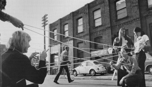 Still Image of Artist Vera Frenkel's String Games: Improvisations for Inter-City Video (Montreal–Toronto, 1974) featuring a street-scene with artist making a cats cradle shape using their bodies and rope 