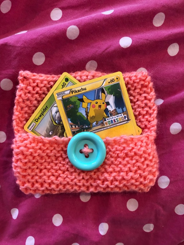 a peachy/pink knitted pouch with a bright blue button. It contains two pokemon cards, one dewpider and one pikachu.