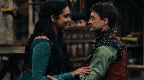 Geraldine Viswanathan and Daniel Radcliffe in medieval comedy Miracle Workers. They are playing Eliza and Chauncely who are just pulling away after a kiss. A villager is watching them covertly from a barrel in the background.