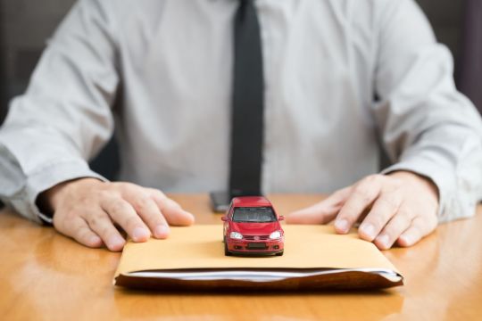 homem de gravata e camisa social segura pacote de documentos com carro vermelho em cima representando indenização de seguro