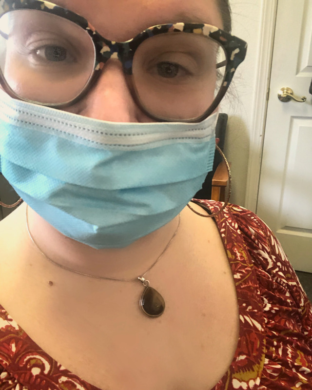 A tired looking woman looks at the camera. She is Caucasian, with her hair pulled back and wearing a surgical mask. she is sitting in an office.