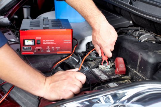 pessoa conecta cabos de um carregador portatil nos terminais da bateria no motor de um carro