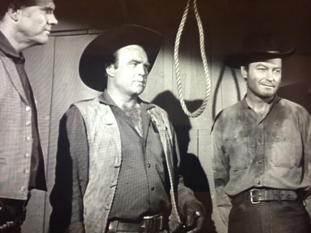 DeForest Kelley stands in a doorway with a very sour puss. equally sour sheriff stands by in profile.