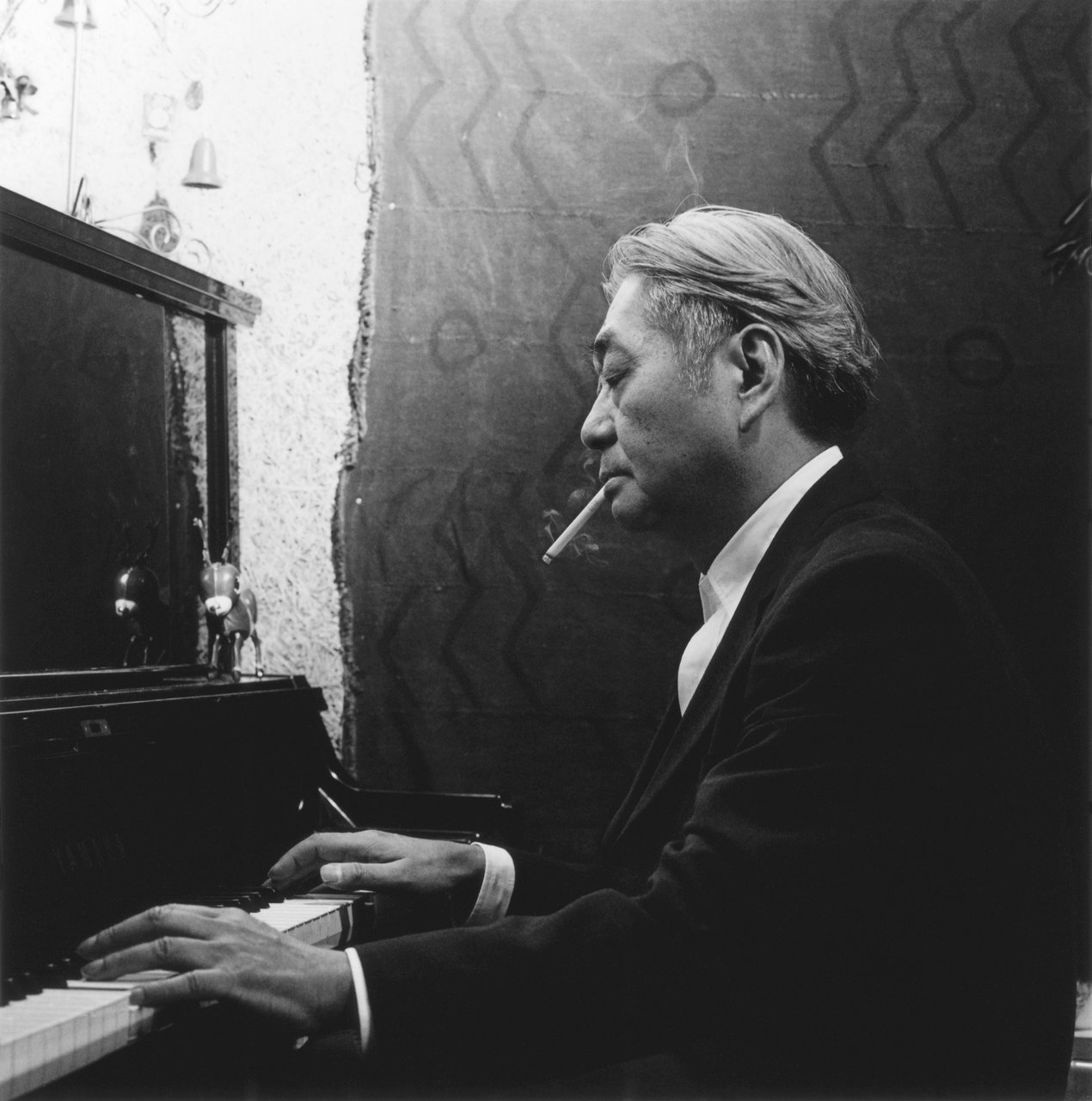 picture of middle age Haruomi Hosono playing a piaco while smoking a cigarette