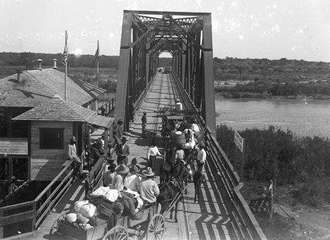 People and wagons crossing a wrought iron bridge over a large river. A building with flags flying over it stands to the left. People are leaning on the bridge railings watching the people crossing.