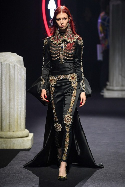 stern looking woman stalks down the runway; her suit is embroidered with a floral pattern that forms the outline of a skeleton
