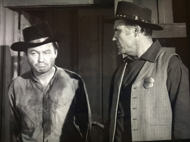DeForest Kelley stands in a doorway with a very sour puss. equally sour sheriff stands by in profile.