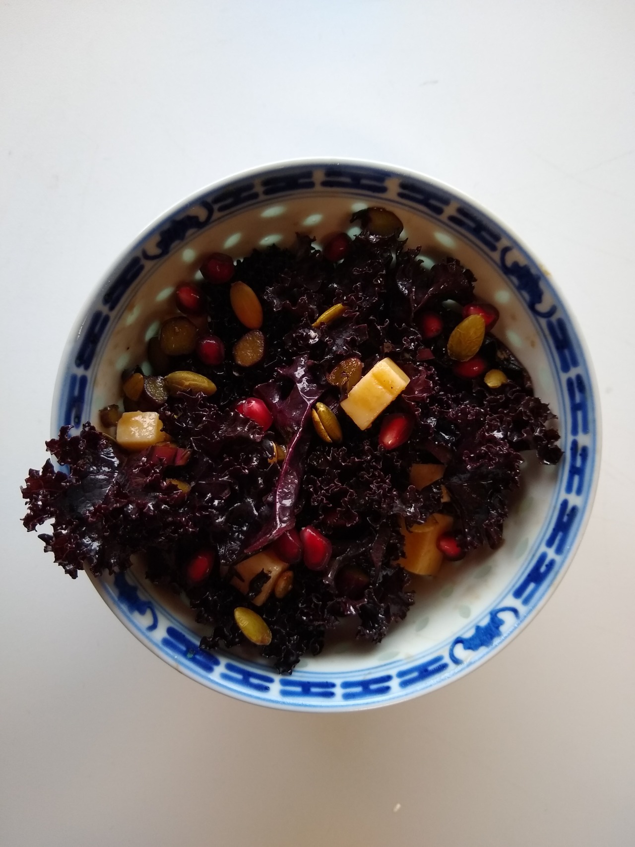 An overhead closeup shot of purple kale salad in a blue and white bowl. The salad has pomegranates, pumpkin seeds, and white cheese in it.