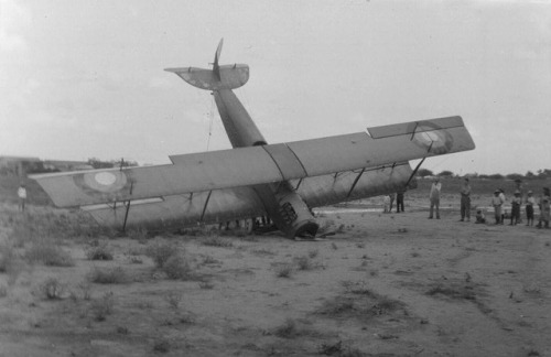 A crashed bi-plane with its nose and one wing touching the ground and it's other wing and tail up in the air. A small group of people stand off to the side. 