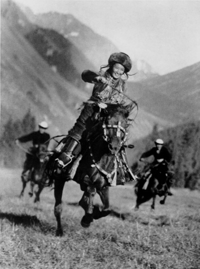 black and white photo of a young woman in a fur hat with braids flying around her face riding a horse, smiling
