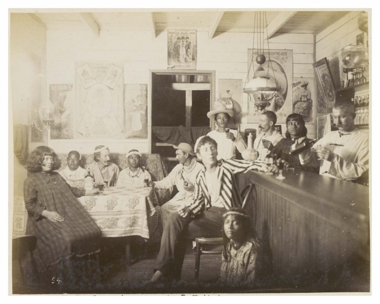 i12bent:A group of people fill the Sans Souci bar-room at Butaritari on the Gilbert Islands. Robert Louis Stevenson sits in between two native women wearing a circlet on his head. Lloyd Osbourne is sitting at bar wearing stripy jacket and holding a...